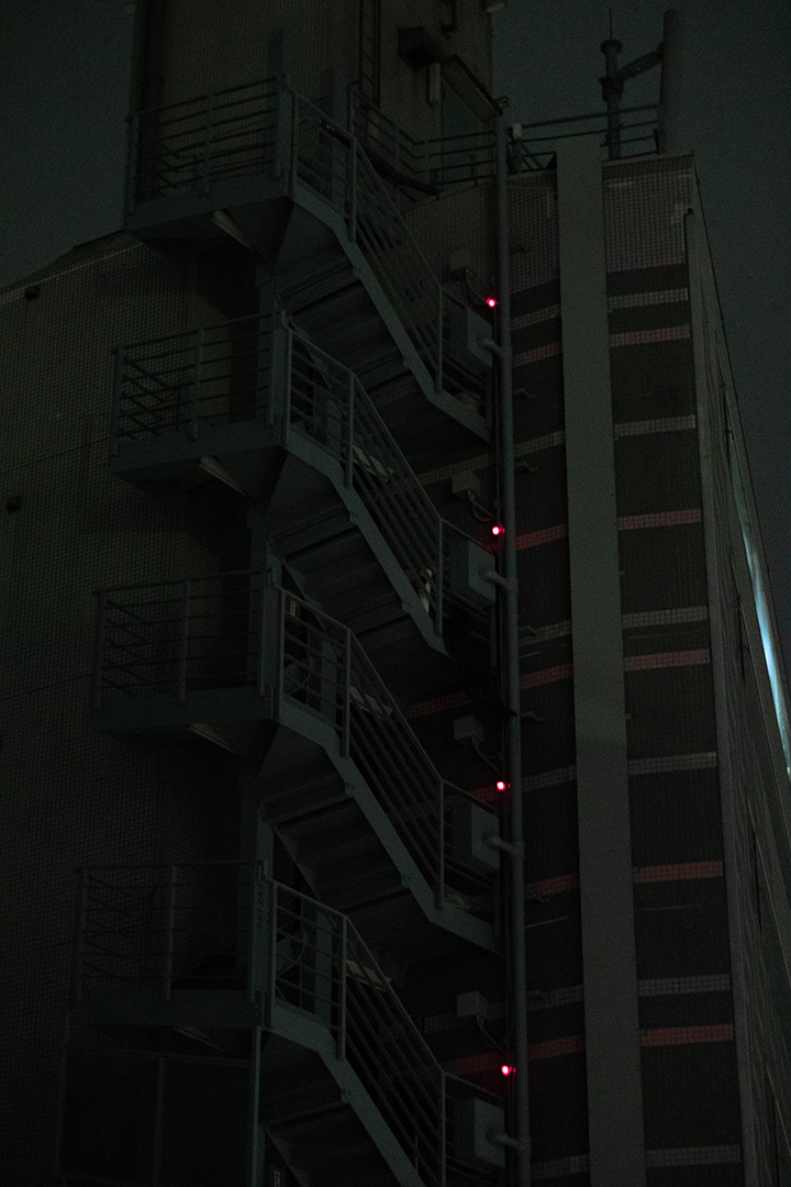Zoom-in to a dark gray building with dark green ceramic tiles and small red lights along its outdoor staircase.